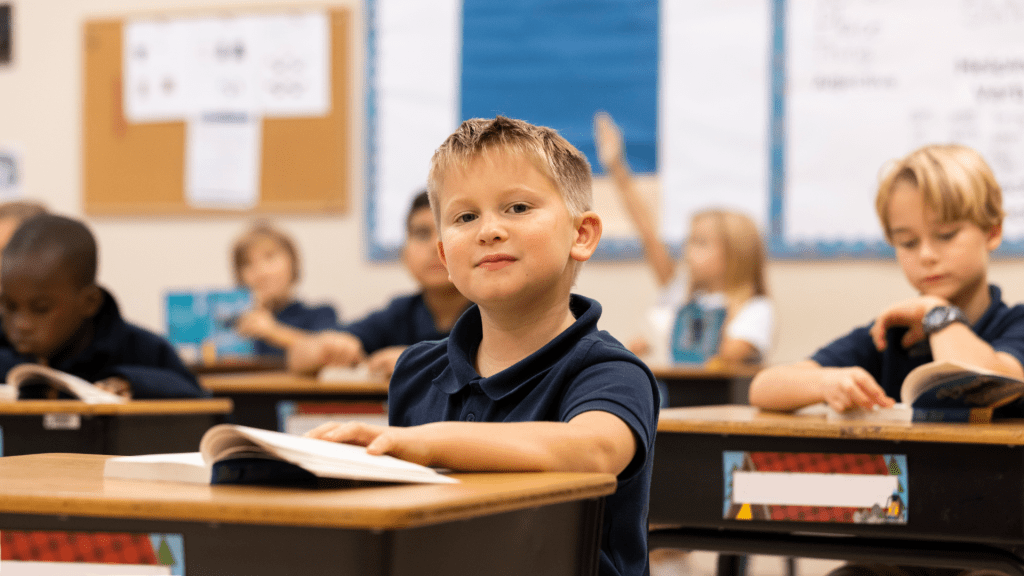 K-3 age boy looks up from his book and into the camera with a thoughtful expression. 