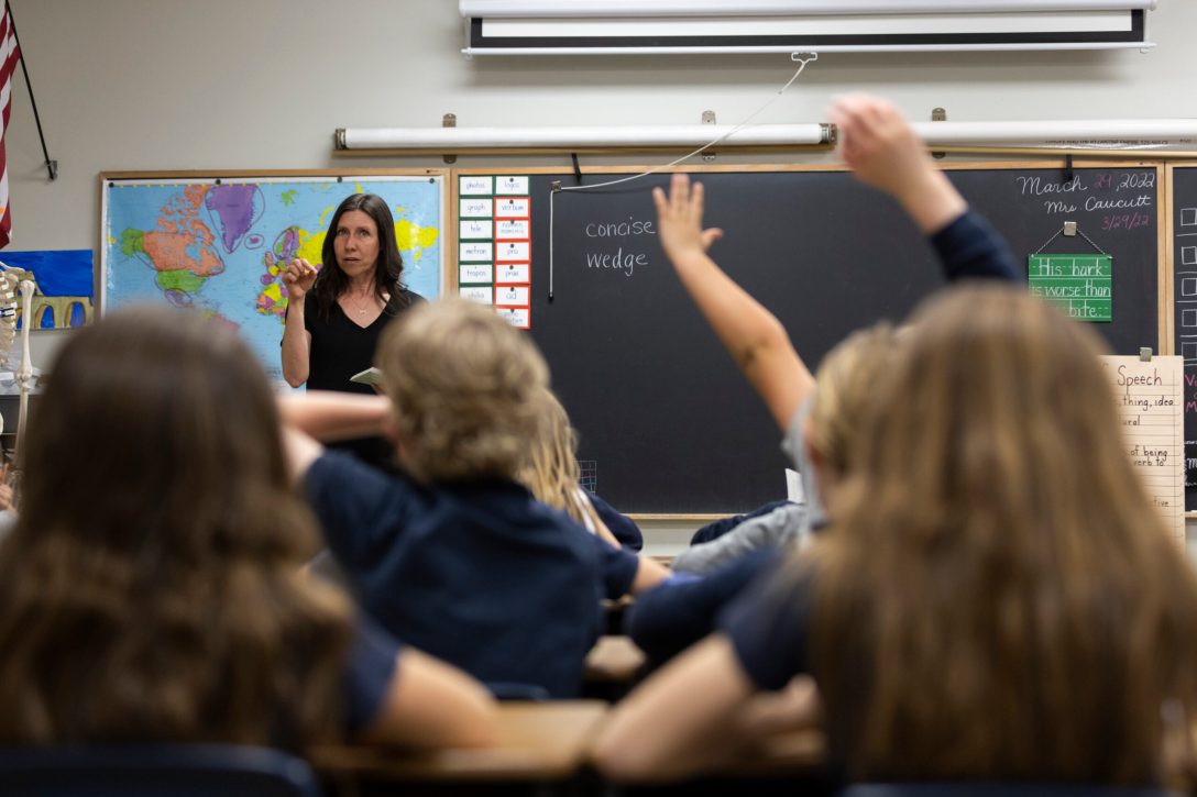 Blurred foreground of students raising their hands with a teacher at the front of the room in focus. A map and a chalkboard can be seen behind her.